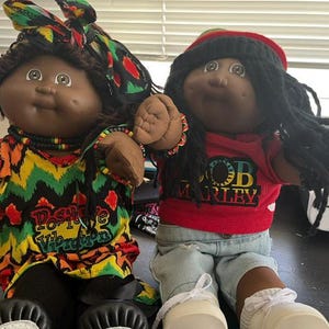Può includere: Due bambole con carnagione scura e capelli di lana. La bambola a sinistra indossa una maglietta colorata con motivo a zigzag con la scritta Positive Vibration e un foulard abbinato. Quella a destra indossa una t-shirt rossa di Bob Marley, pantaloncini di jeans e un berretto.