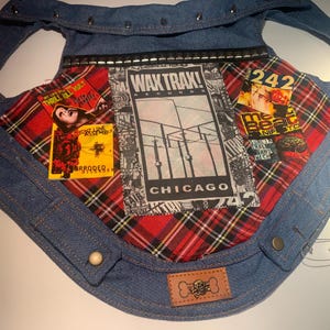 May include: A denim dog vest with a red tartan lining. The vest features patches with band logos and the text "WAX TRAX! RECORDS CHICAGO". The vest has metal studs and a brown leather patch with a skull and crossbones.