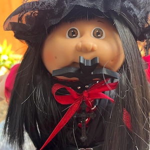 Könnte beinhalten: Puppe mit hellem Teint, braunen Augen und langem schwarzem Haar. Die Puppe trägt ein schwarzes Kleid mit roter Schleife und Kreuzanhänger. Ein schwarzer Spitzenhut ziert den Kopf der Puppe und erzeugt eine gotische Ästhetik.