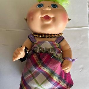 May include: A doll with bright neon green hair, blue eyes, and rosy cheeks. It wears a pink, white, and purple plaid dress with a metallic studded belt, a studded choker necklace, a black studded wristband, and a purple glittery wrist accessory.