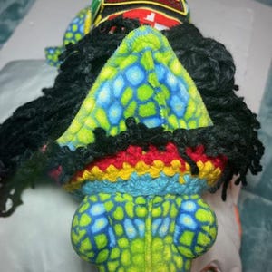 May include: A plush toy with a green, blue, and yellow patterned head and body, black dreadlocks, and a red, yellow, and blue crocheted band. The toy has a tag that reads "One World".