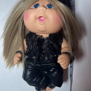 May include: A Cabbage Patch Kid doll with blonde hair, blue eyes, and rosy cheeks, wearing a black leather-look outfit, including a cap, vest, and pants. The doll has a playful expression with its tongue sticking out. The doll is approximately 8 inches tall.