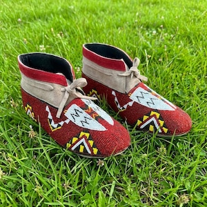 May include: A pair of red beaded moccasins with intricate white, yellow, and black geometric patterns. The ankle boots have tan suede accents, red trim, and tan laces. The shoes are displayed on a bed of green grass.