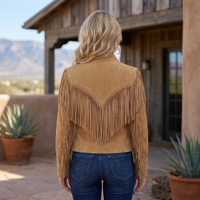 May include: A tan suede jacket featuring long fringe detailing across the back and sleeves, worn with dark blue denim jeans. The jacket has a V-shaped yoke design on the back, with the fringe extending from the shoulders down to the mid-back.