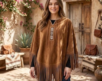 May include: A tan suede poncho featuring long fringe trim along the hemline and a central rectangular panel with a geometric pattern. The garment has a V-neckline with thin decorative ties and is worn over a black long-sleeved shirt and blue jeans.