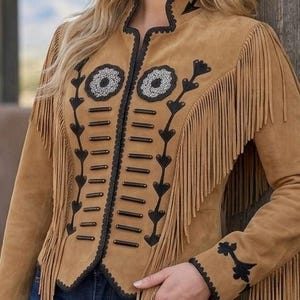 May include: A tan suede jacket featuring long fringe detailing along the sleeves and chest. The front is adorned with black decorative trim, horizontal bar accents, and two circular floral-inspired embellishments with silver-toned beadwork.