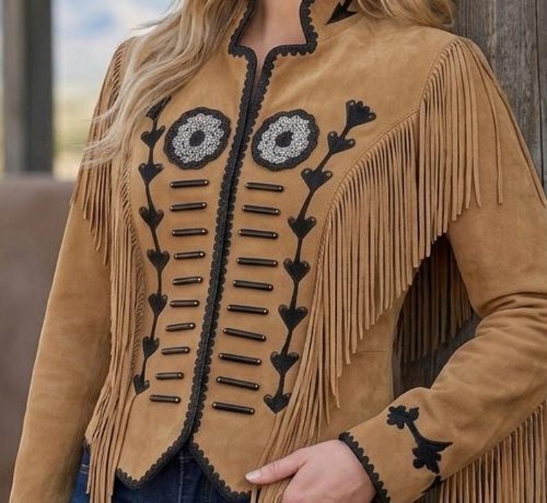 May include: A tan suede jacket featuring long fringe detailing along the sleeves and chest. The front is adorned with black decorative trim, horizontal bar accents, and two circular floral-inspired embellishments with silver-toned beadwork.