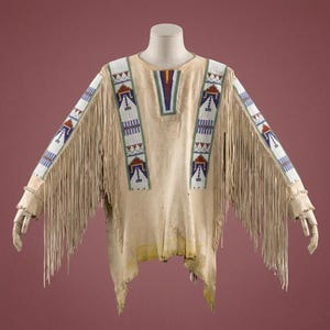 May include: A beige leather shirt featuring long fringe along the sleeves and sides. The garment is decorated with vertical panels of white, blue, red, and green beadwork on the chest and sleeves, showcasing geometric patterns and designs.