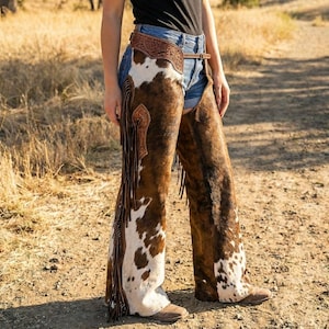 Peut inclure: Chaps en peau de vache avec un motif tacheté marron et blanc, avec des accents décoratifs en cuir et des franges. Les chaps sont portés avec une ceinture en cuir marron. Un article de style western classique.