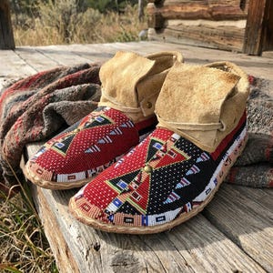 May include: A pair of tan suede ankle boots featuring intricate beadwork on the vamps. The beadwork displays geometric patterns in red, black, white, and blue. The boots have a fold-over cuff design and are secured with a small brass-colored stud.