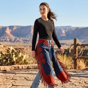 May include: A long-sleeved black crewneck shirt worn with blue denim jeans and suede chaps. The chaps feature a deep blue center panel with red floral appliqu&eacute;s, red suede trim along the edges, and long fringe detailing hanging from the sides.