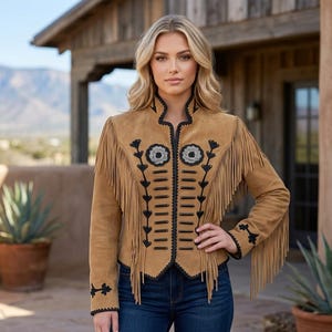 May include: A tan suede jacket featuring long fringe detailing along the chest and sleeves, accented with black embroidery and decorative black cord trim down the front, paired with dark blue denim jeans.
