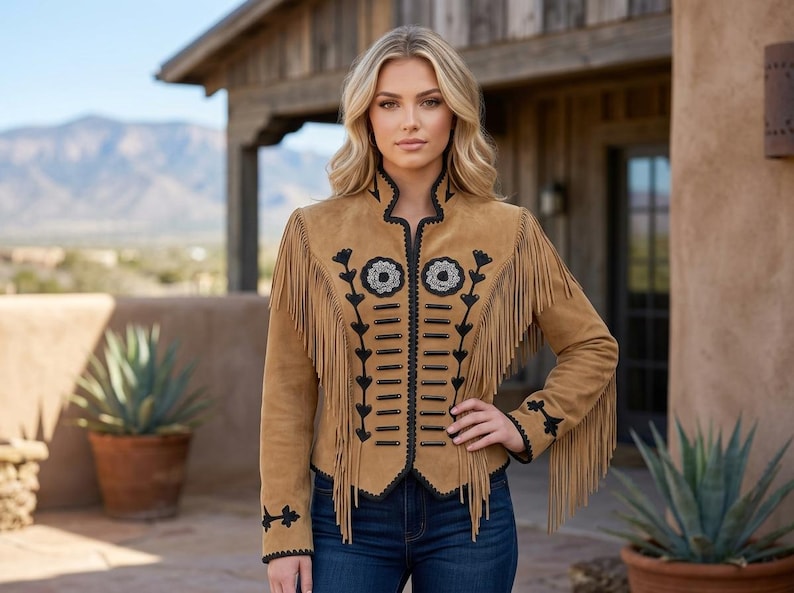 May include: A tan suede jacket featuring long fringe detailing along the chest and sleeves, accented with black embroidery and decorative black cord trim down the front, paired with dark blue denim jeans.