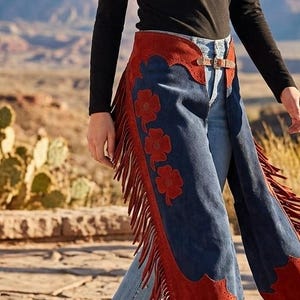 May include: Blue suede chaps featuring long red fringe along the outer legs and decorative red floral cutouts. The chaps have a red suede waistband with a metal buckle closure at the front, worn over light blue denim jeans and a black long-sleeve top.