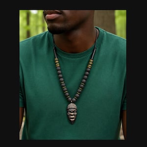 May include: A necklace featuring a dark metallic pendant shaped like a stylized human face with textured hair. The cord is strung with matte black spherical beads interspersed with small, weathered green and gold cylindrical spacers.