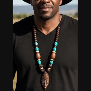 May include: A long necklace featuring a central carved wooden mask pendant, accented with alternating dark brown wooden beads, smooth turquoise-colored beads, and small cream-colored spacers, all strung on a thin black cord against a black shirt.