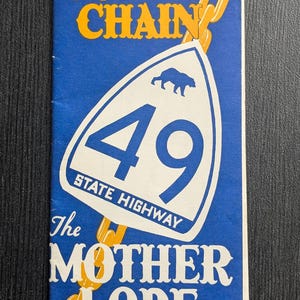 May include: A vintage travel brochure for California State Highway 49. The blue and white sign features a bear and the number 49. The brochure's text includes "California's Golden Chain" and "The Mother Lode Highway" in gold lettering.