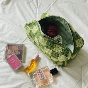 May include: A green and white checked crochet cosmetic bag with a floral lining, containing beauty products. Includes a Dior eyeshadow palette, Dior blush, perfume, and a small bottle of oil.