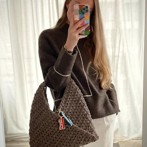 Peut inclure: Un sac à main marron en crochet de forme triangulaire. Le sac est orné d'un porte-clés avec un charm en forme de lettre "E" rouge et blanc. La personne porte un pull marron et un pantalon blanc.