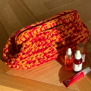 May include: A red and orange crocheted bag with three small bottles of orange liquid and a tube of cream with the text "LIERACTY" and "VICHY" on a wooden surface.