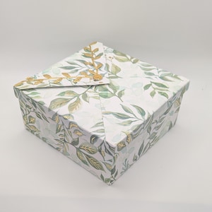 May include: Square gift box with a white background. The box is decorated with a watercolor pattern of green leaves and gold accents. The lid is folded over and secured with a gold leaf design.