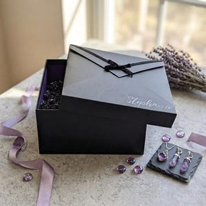 May include: A square black gift box with a black ribbon bow on the lid, featuring the name Stephanie in white script. The box is partially open, revealing dark filler material inside. Three purple crystal pendants sit on a small black slate tile.