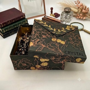May include: A square black box with a copper-colored floral line art pattern and the word CRYSTAL printed on the lid. The lid features a gold-colored branch embellishment and a circular seal. The box is partially open, revealing dark contents inside.