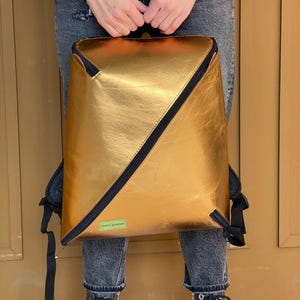 May include: A gold metallic backpack with black straps and a black zipper. The backpack has a diagonal seam across the front.