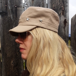 May include: A woman with long blonde hair is wearing a brown cap with silver star embellishments. The cap has a curved brim and a structured crown. She is wearing a brown sweater.