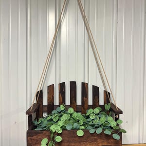 Puede incluir: Una jardinera colgante de madera con acabado marrón oscuro, llena de plantas verdes con hojas. La jardinera tiene respaldo de listones y está suspendida por una cuerda natural. El diseño se asemeja a un pequeño banco.