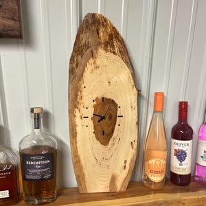 May include: A rustic wooden clock with a natural edge, featuring a light brown color and visible wood grain. The clock face has black hands and minimalist hour markers. Bottles of liquor and wine are on a wooden shelf below the clock.