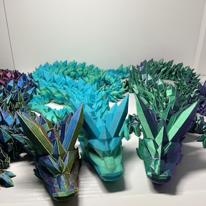 May include: Three articulated dragon sculptures in shades of blue, green, and purple. Each dragon is made of geometric shapes that resemble crystals. The dragons are lying on a white surface.