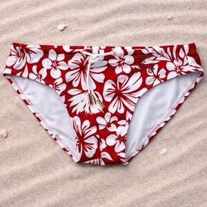 Pode incluir: Cueca de banho com estampa floral vermelha e branca com cintura com cordão. O traje de banho é feito de um material leve e foi projetado para nadar. A cueca é mostrada em uma superfície arenosa.