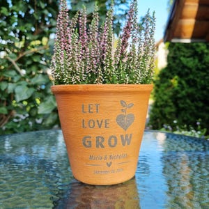 May include: A terracotta-colored flower pot with the words "LET LOVE GROW" and a heart-shaped plant design. The pot contains a flowering plant with pink and green foliage. The names "Maria & Nicholas" and the date "September 20, 2025" are also printed on the pot.