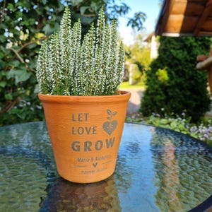 May include: A terracotta-colored flower pot with the words "LET LOVE GROW" and a heart-shaped design. The pot contains a plant with green stems and white flowers. The names "Maria & Nicholas" and the date "September 20, 2015" are also printed on the pot.