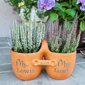 Peut inclure: Un pot de fleurs en terre cuite avec deux pots reliés, chacun contenant des plantes à fleurs. Le pot de gauche porte l'inscription "Mr. Lewis" et le pot de droite "Mrs. Lewis" avec la date "10.17.25" au milieu. Les plantes ont des fleurs blanches et roses.