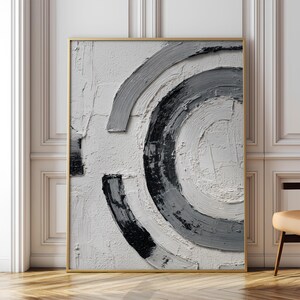 Peut inclure: Tableau abstrait avec un cadre doré. L'œuvre présente un fond blanc texturé avec des formes courbes grises et noires qui se chevauchent. La peinture est exposée dans une pièce avec des boiseries blanches et un sol en bois.
