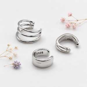 May include: Three silver-coloured ear cuffs with a polished finish. One cuff features a triple-band design, while the other two are single-band styles. The two single-band cuffs have the text S925 clearly stamped on their inner surfaces.