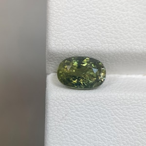 May include: An oval-cut, olive-green gemstone with facets. The gem has a mix of light and dark green hues, with a slightly translucent appearance. The stone is set against a white background.