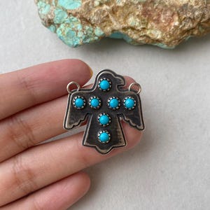 May include: A silver-coloured pendant in the shape of a stylised bird, decorated with seven round, bright turquoise stones. The pendant has a weathered, antique finish and includes two small loops for hanging.
