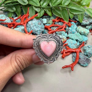 Könnte beinhalten: Ein silberner Ring mit einem rosa herzförmigen Stein. Der Ring hat ein aufwendiges, detailliertes Design um das Herz und am Band. Der Ring wird vor einem Hintergrund aus türkisfarbenen Steinen und roter Koralle gezeigt.