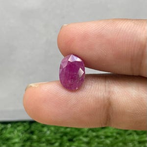 May include: A faceted, oval-shaped ruby gemstone. The ruby is a deep, vibrant red color, with multiple facets that catch the light. The gemstone is held between two fingers against a blurred background.