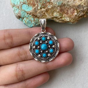 May include: A silver pendant with a circular design, featuring turquoise stones and a detailed border. The pendant has a bail for a chain. The turquoise stones are arranged in a pattern, with a larger stone in the center.