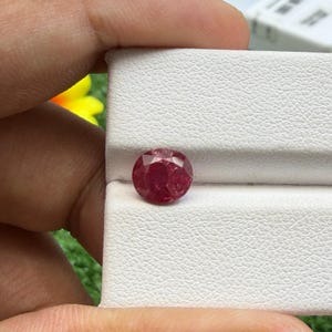 May include: A round, faceted, deep red gemstone. The gem is displayed in a white tray, with a hand partially visible. The stone's facets reflect light, showcasing its brilliance. The background includes a glimpse of a yellow flower and green foliage.