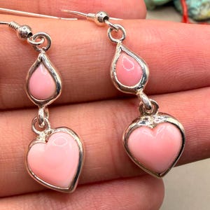May include: A pair of drop earrings featuring polished pink heart-shaped and teardrop-shaped stones set in silver-toned metal. Each earring consists of a small teardrop stone suspended above a larger heart-shaped stone, attached to a silver hook.