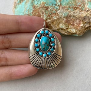 May include: Silver-toned pendant with a teardrop shape, featuring a central oval turquoise stone surrounded by smaller turquoise stones. The pendant has a sunburst design at the bottom.
