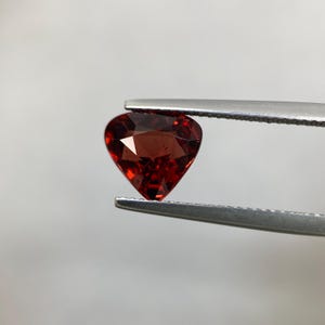 May include: A heart-shaped, faceted, deep red gemstone is held by a pair of silver tweezers. The gemstone's facets reflect light, creating a sparkling effect. The background is a soft, neutral gray.