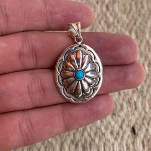 May include: Silver-toned pendant with an oval shape, featuring a detailed floral design. The center of the flower has a small, round, turquoise-colored stone. The pendant has a bail at the top for attaching to a chain.