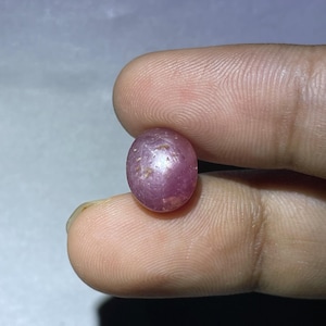 Może przedstawiać: Okrągły, różowy rubin z gwiaździstym wzorem. Kamień szlachetny ma około 1 cm średnicy.