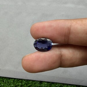 May include: A faceted, oval-shaped, deep blue gemstone. The gem is held between fingers, showcasing its polished surface and color. The stone's facets catch the light, creating a sparkling effect. This gemstone is suitable for jewelry making.